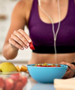 Mulher com roupa de desporto roxa e auriculares, numa cozinha, a preparar uma taça azul com fruta, colocando um pedaço de morango por cima. Na mesa estão também outras frutas cortadas e inteiras.
