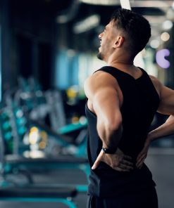 Homem de costas, com roupa de treino preta, num ginásio, com as mãos na zona lombar e expressão de desconforto, rodeado por equipamentos de musculação desfocados ao fundo.