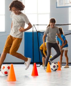 Crianças a realizar exercícios de condução de bola com obstáculos em interior, usando bolas de futebol e cones coloridos, num espaço desportivo com baliza ao fundo.