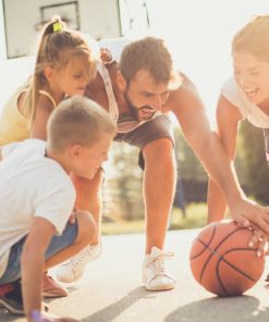 Família a brincar com uma bola de basquetebol num campo exterior, com expressões de alegria e envolvimento de todos os elementos.