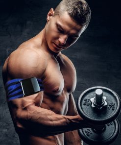 Homem musculado sem camisola a segurar um haltere com o braço direito, usando uma braçadeira de oclusão no braço, num ambiente escuro com fundo neutro.