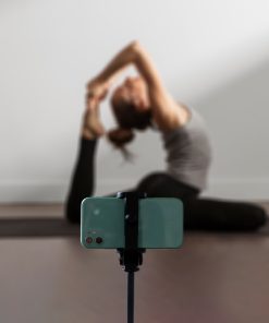Smartphone montado num tripé em primeiro plano, com uma mulher ao fundo a praticar yoga numa sala, numa posição de alongamento com uma perna esticada e as mãos a segurar o pé por trás da cabeça.