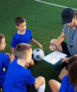 Grupo de crianças com equipamento desportivo azul sentadas à volta de um adulto com apito ao pescoço, que segura uma prancheta com um esquema tático desenhado, num campo de relva sintética.