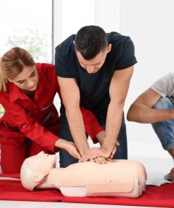 Homem a realizar compressões torácicas num manequim de reanimação, sob orientação de uma formadora, enquanto outros participantes observam atentamente durante uma sessão de primeiros socorros.