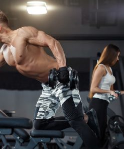 Homem sem camisa realiza exercício de remada unilateral com haltere, apoiado num banco de treino; ao fundo, mulher treina com halteres junto ao suporte.