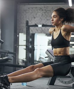Mulher a realizar exercícios de tríceps num banco de musculação, vestida com roupa desportiva preta, num ginásio equipado com máquinas de musculação ao fundo.