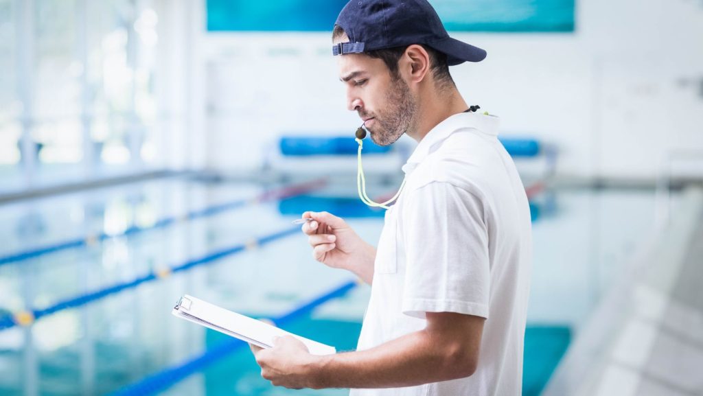 Treinador de natação na piscina com apito e bloco de notas, em ambiente aquático interior, focado na segurança, desempenho e organização de treinos.