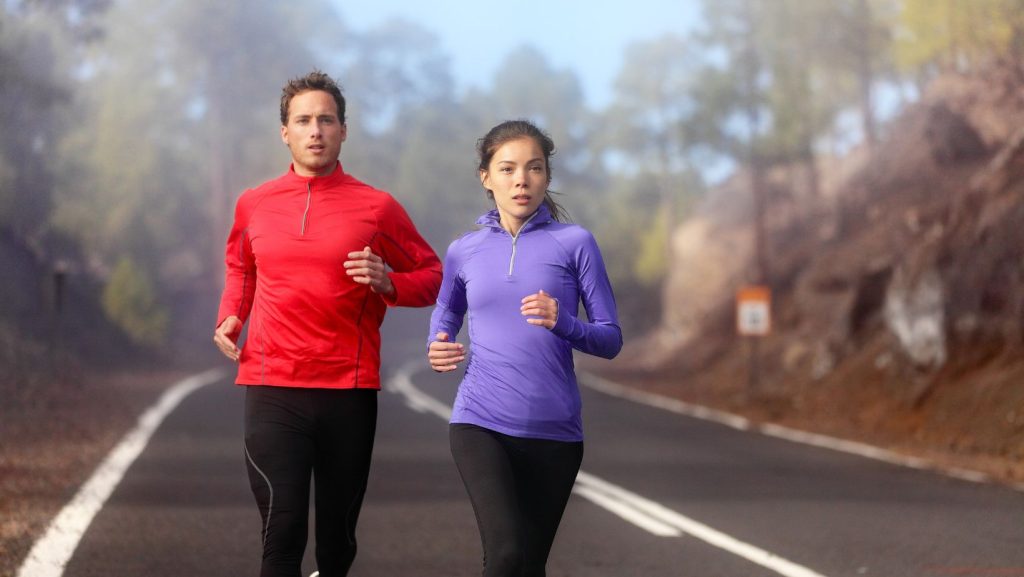 Casal jovem a correr numa estrada montanhosa com neblina, usando roupa desportiva de inverno, símbolo de saúde, motivação e estilo de vida activo.