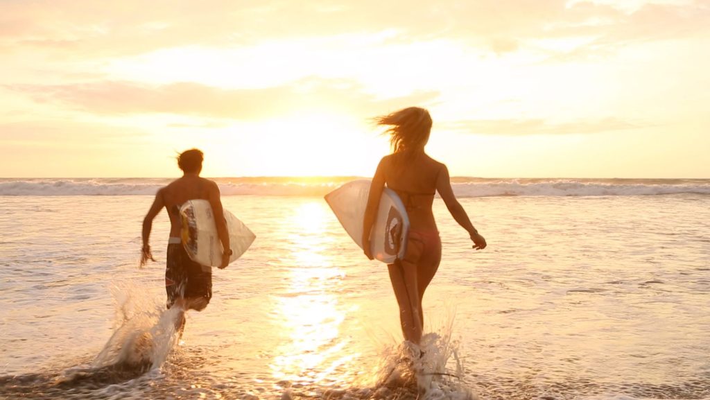 Dois surfistas, um homem e uma mulher, entram no mar ao pôr do sol, com pranchas na mão, num cenário de liberdade, aventura e conexão com a natureza.