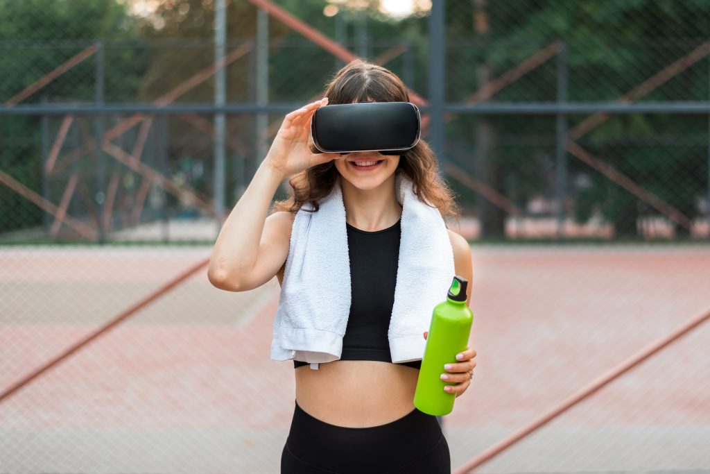 Mulher jovem com garrafa de água e toalha ao pescoço, a usar óculos de realidade virtual num ambiente desportivo, simbolizando inovação no fitness.