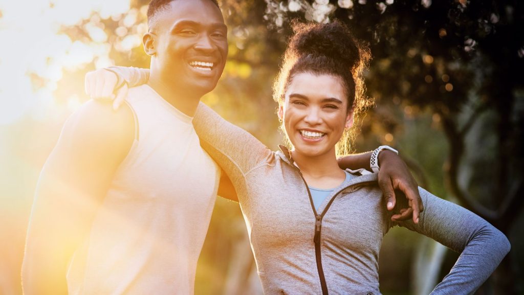 Casal jovem a sorrir ao ar livre durante o pôr do sol, com roupa desportiva, transmitindo bem-estar, amizade e estilo de vida saudável ao praticar desporto.