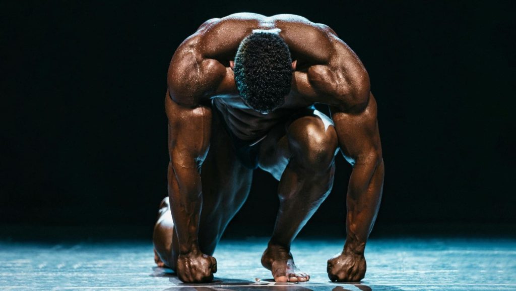 Físico masculino musculado ajoelhado em pose poderosa sob luz dramática, simbolizando força, disciplina e superação no culturismo.