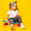 Menina sentada num skate azul com rodas cor-de-rosa, sobre fundo amarelo vivo. Usa calças de ganga, t-shirt branca, ténis claros e uma bandolete azul.