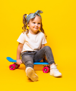 Menina sentada num skate azul com rodas cor-de-rosa, sobre fundo amarelo vivo. Usa calças de ganga, t-shirt branca, ténis claros e uma bandolete azul.