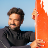 Homem de fato de neoprene segura uma prancha de surf vermelha, com o mar e céu azul ao fundo.