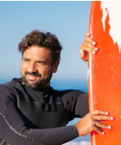 Homem de fato de neoprene segura uma prancha de surf vermelha, com o mar e céu azul ao fundo.